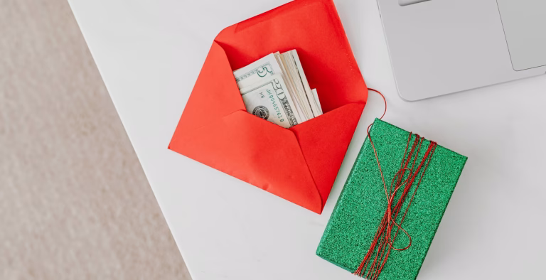 A green gift box and red envelope with cash on a desk beside a laptop, emphasizing holiday or financial themes.